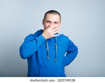 Portrait Of A Man Closes His Nose, Smells Bad, Dressed In A Blue Hoodie, Isolated On A Gray Background