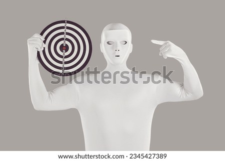 Portrait of a man disguised in a white spandex bodysuit and wearing a white mask pointing at a round goal board target that he is holding in his hand isolated on a grey background. Goal concept