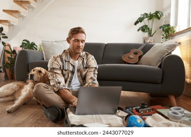 Portrait of man with dog, sitting on floor, has suitcase and laptop opened, booking or buying online, planning holiday, packing for vacation. - Powered by Shutterstock