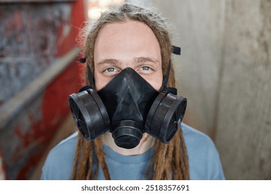 Portrait of man with dreadlocks looking at camera while wearing industrial respirator mask in urban setting. Background includes graffiti on metal and concrete walls - Powered by Shutterstock