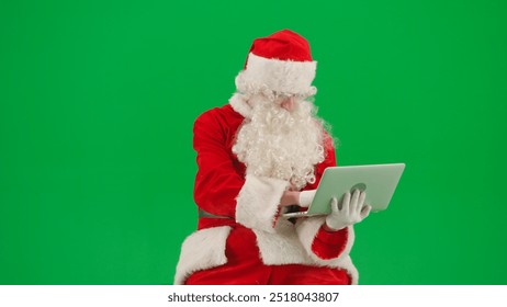 Portrait of man dressed as Santa on chroma key green screen background. Santa Claus in outfit sitting holding laptop typing, messaging online. - Powered by Shutterstock