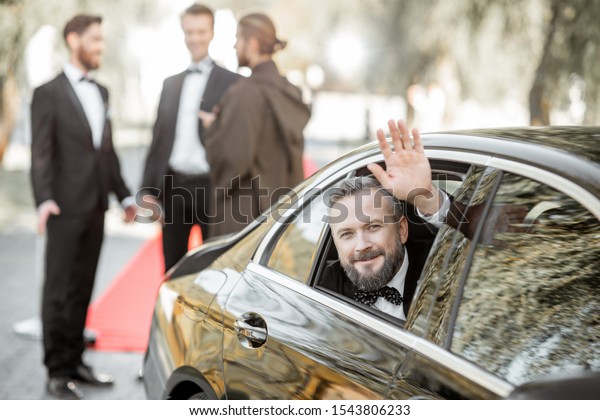 Portrait of a man as a famous movie actor
sitting in the luxury car, arriving on the awards ceremony or movie
premiere near the red carpet
outdoors