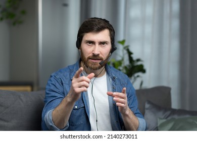 Portrait of a man with a headset working online consultant at home, focused and angry talking to a client, looking at the webcam - Shutterstock ID 2140273011
