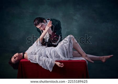Portrait of man in image of vampire biting hand of young woman lying on table over green background. Blood drinking. Concept of Halloween, mystery, gothic art, fantasy, surreal characters