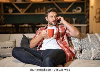 Portrait of man looking at smart phone at living room. Man is talking hold cup of coffe chatting on the smartphone. Thoughtful man using mobile phone at home. Middle-aged man using phone. - Powered by Shutterstock