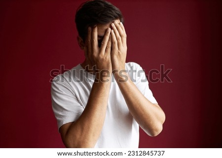 Portrait of man looking thorough fingers covering face with palms, feeling shy and embarrassed standing against red studio background. People and emotions, signs, gestures, body language concept