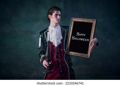 Portrait Of Man In Noble Medieval Costume In Image Of Vampire Holding Picture Frame Isolated Over Dark Green Background. Hunter. Concept Of Halloween, Mystery, Gothic Art, Fantasy, Surreal Characters