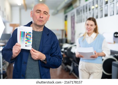 Portrait Of Man Printshop Worker Holding Colour Test Page For Printer. Woman Manager Standing In Background.