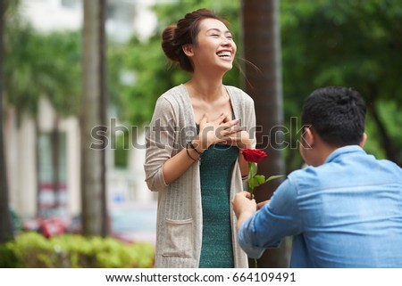 Portrait of man proposing to smiling girlfriend in street, standing on one knee and holding red rose