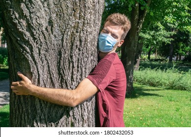 Portrait Of Man In Protective Face Mask Hugging Tree Trunk In Park. Man Wearing Medical Mask Embracing Wood Trunk. Longing For Hugs. Human Connection To Nature Through Tree