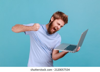 Portrait Of Man Punching Laptop Screen, Looking With Furious Mad Expression, Boxing Threatening To Hit While Having Online Conversation, Video Call. Indoor Studio Shot Isolated On Blue Background.