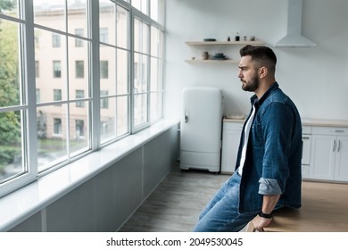 Portrait of man sad and depressed in despair at home, covid-19 lockdown, agoraphobia, personal problems. Unhappy millennial european handsome man looks at large window in kitchen interior, profile
