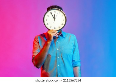 Portrait of man in shirt hiding face behind big wall clock display, wasting his time, procrastination, bad organization of working time. Indoor studio shot isolated on colorful neon light background. - Shutterstock ID 2115567854