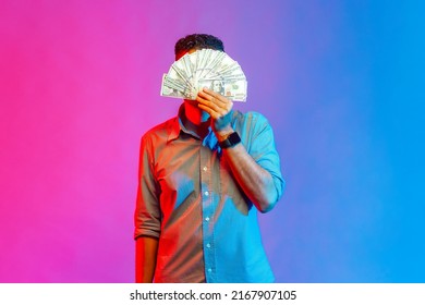 Portrait of man in shirt hiding face behind bunch of dollar banknotes, anonymous person holding money, lottery win, big profit. Indoor studio shot isolated on colorful neon light background.