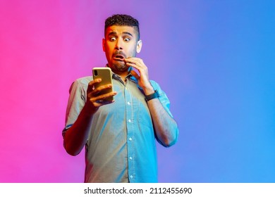 Portrait of man in shirt looking at smartphone display with funny scared face, shocked and surprised with application. Indoor studio shot isolated on colorful neon light background.