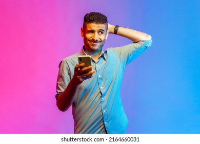 Portrait of man in shirt scratching head holding smartphone in hand, contemplating about software updating, choosing suitable tariffs. Indoor studio shot isolated on colorful neon light background. - Shutterstock ID 2164660031