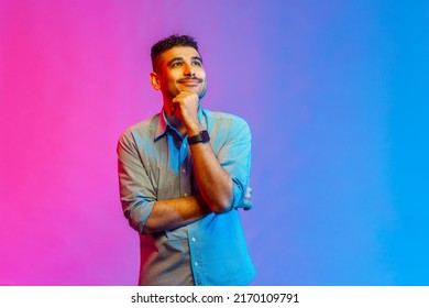 Portrait of man in shirt standing, touching his chin, toothy smile and looking away dreaming, imagining and fantasizing. Indoor studio shot isolated on colorful neon light background.