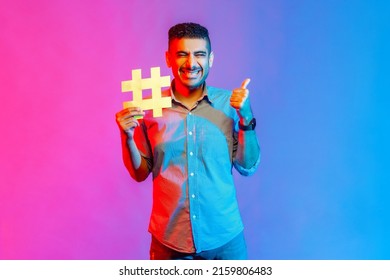 Portrait of man in shirt with toothy smile, holding hashtag symbol, showing thumb up, recommending popular topics, internet trends. Indoor studio shot isolated on colorful neon light background.