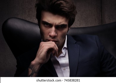 Portrait Of A Man Sitting In A Chair. Sinister Look. White Shirt And Dark Blue Jacket