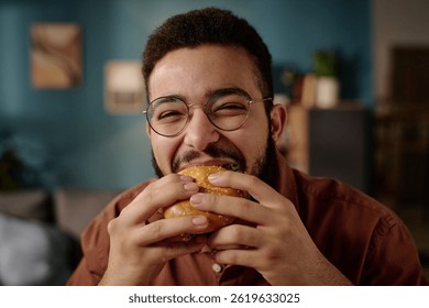 Portrait of man smiling warmly while eating a delicious burger indoors in cozy, modern setting with framed pictures on wall in background