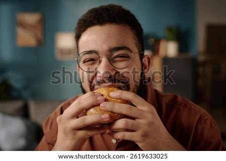 Portrait of man smiling warmly while eating a delicious burger indoors in cozy, modern setting with framed pictures on wall in background