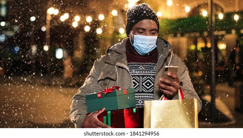 Portrait of man standing and texting on mobile phone. Young African American happy male with little Christmas tree and shopping bags typing on smartphone while snowing. Winter holidays concept