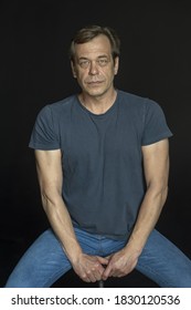 Portrait Of A Man In A Studio 40-50 Years Old In A Blue T-shirt With Strong Hands On A Black Blurry Background. Maybe He's Just A Buyer, An Actor Or A Truck Driver, A Loader Or A Military Pensioner,