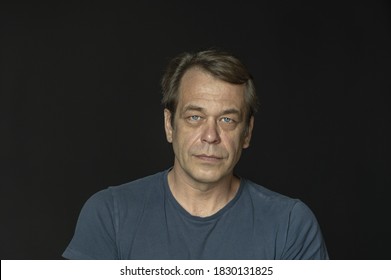 Portrait Of A Man In A Studio 40-50 Years Old In A Blue T-shirt On A Black Blurry Background, Close-up. Maybe He's Just A Buyer, An Actor Or A Truck Driver, A Loader Or A Military Pensioner