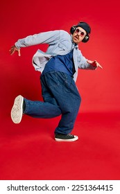 Portrait of man in stylish clothes, oversized jeans, shirt and hat posing, dancing over red background. Concept of modern fashion, lifestyle, music culture, emotions, facial expression. Ad