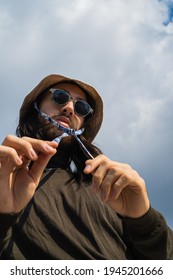 Portrait Of A Man With Sunglasses And A Bucket Hat. Looking Confidently At The Camera. Blue Sky In The Background. Fashion Photography. Low Angle View.