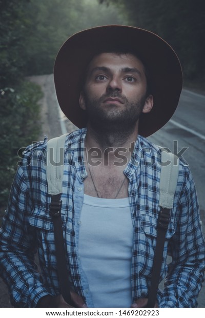 Portrait: Man Travel blogger in\
velvet hat and hiking backpack stands in a foggy road in the\
forest.