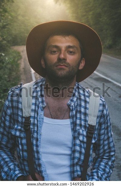 Portrait: Man Travel blogger in\
velvet hat and hiking backpack stands in a foggy road in the\
forest.