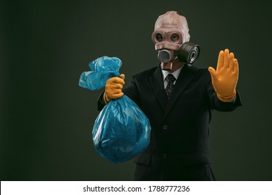 Portrait Of A Man Wearing A Dark Business Suit And An Old Gas Mask Holding A Blue Garbage Bag And Showing Stop, Isolated On Dark Green Background.