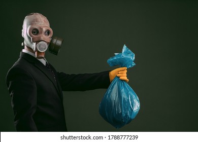 Portrait Of A Man Wearing A Dark Business Suit And An Old Gas Mask Holding A Blue Garbage Bag, Isolated On Dark Green Background.