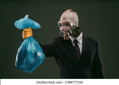 Portrait Of A Man Wearing A Dark Business Suit And An Old Gas Mask Holding A Blue Garbage Bag, Isolated On Dark Green Background.