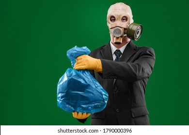 Portrait Of A Man Wearing A Dark Business Suit And An Old Gas Mask Holding A Blue Garbage Bag, Isolated On Green Background.