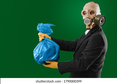 Portrait Of A Man Wearing A Dark Business Suit And An Old Gas Mask Holding A Blue Garbage Bag, Isolated On Green Background.