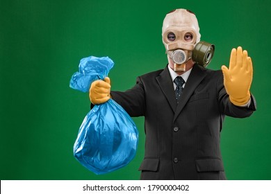 Portrait Of A Man Wearing A Dark Business Suit And An Old Gas Mask Holding A Blue Garbage Bag And Showing Stop, Isolated On Green Background.