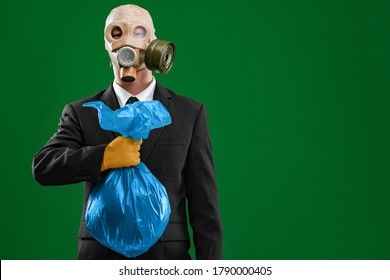 Portrait Of A Man Wearing A Dark Business Suit And An Old Gas Mask Holding A Blue Garbage Bag, Isolated On Green Background.