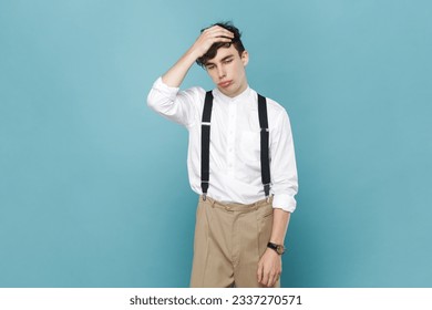 Portrait of man wearing white shirt and suspender standing with facepalm gesture, blaming himself, feeling regret because of bad memory. Indoor studio shot isolated on blue background.
