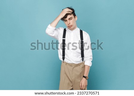 Portrait of man wearing white shirt and suspender standing with facepalm gesture, blaming himself, feeling regret because of bad memory. Indoor studio shot isolated on blue background.