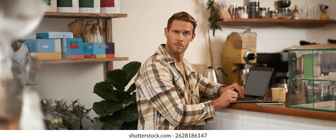 Portrait of man working in cafe, looking confused at camera, sitting with laptop and glass of coffee, staring puzzled at you.