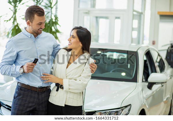 portrait of married\
caucasian couple in car showroom. young man and woman stand posing\
against the backdrop of a new car, look at each other and hold keys\
from car in hands