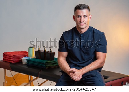 Portrait of a masseur. Masseur grooves into couches. Massage couch is behind him. Massage supplies in background. Masseur looks into camera. Concept of work in spa area. Portrait manual therapist.