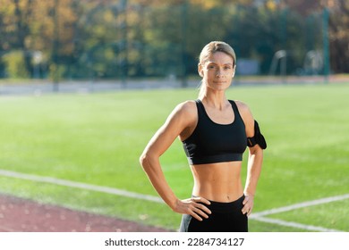 Portrait of mature adult female athlete in stadium, female athlete holding hands on hips smiling and looking at camera, jogging woman in tracksuit during active exercise and fitness class.