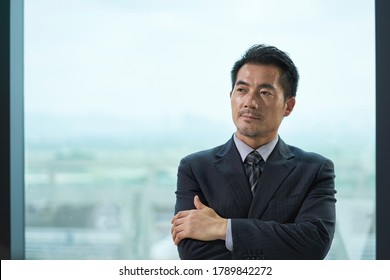 Portrait Of A Mature Asian Business Man Standing By Window In Office Arms Crossed