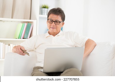 Portrait Of Mature Asian Man Using Computer Doing Online Shopping With Credit Card, Sitting On Sofa At Home, Senior Retiree Indoors Living Lifestyle. 