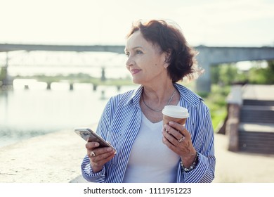 Portrait of a mature attractive stylish woman retired with a cup of coffee and a smartphone in her hands outdoors on a summer sunny day - Powered by Shutterstock