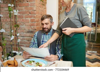 Portrait of mature bearded man reading menu in modern cafe with young waitress giving him ordering advice on business lunch - Powered by Shutterstock