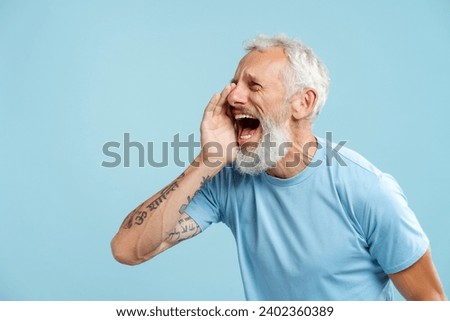 Portrait of mature bearded man shouting announcing something, looking away wearing stylish casual blue t shirt standing isolated on blue background copy space. Communication concept